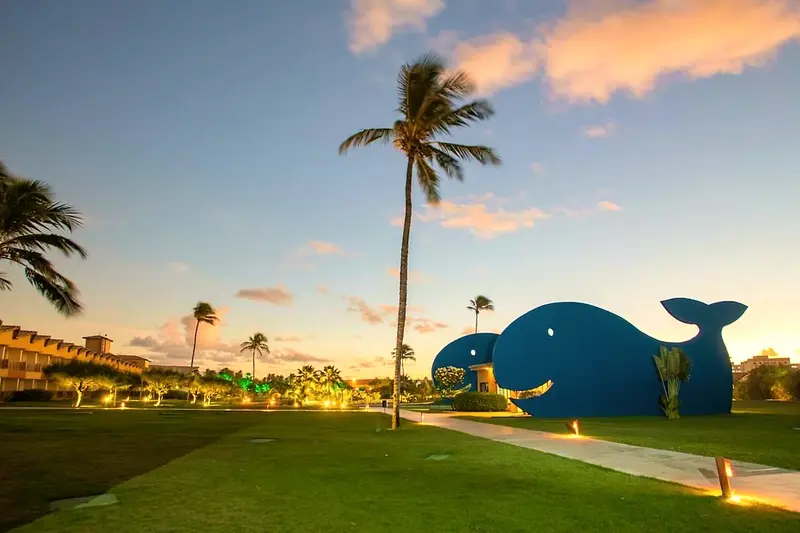 Escultura de baleia azul no gramado iluminado ao entardecer, entre coqueiros