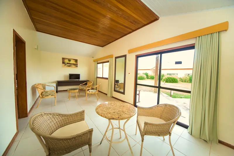 Sala do bangalô com mesa redonda, poltronas de vime e porta de vidro para a varanda