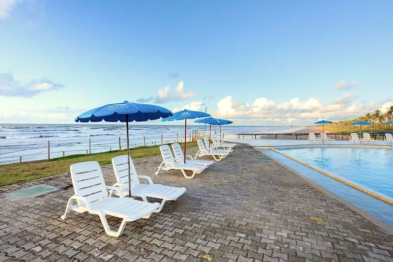 Espreguiçadeiras brancas e guarda-sóis azuis à beira da piscina, de frente para o mar