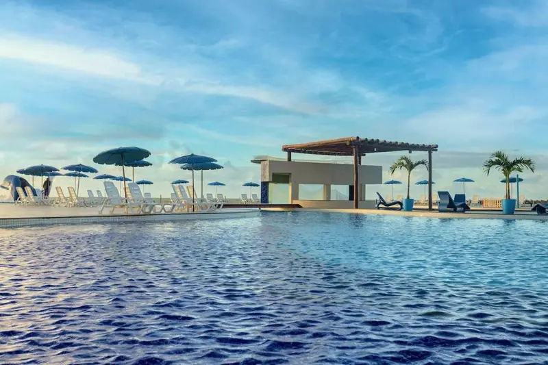 Piscina com bar coberto ao fundo e guarda-sóis enfileirados à beira da água
