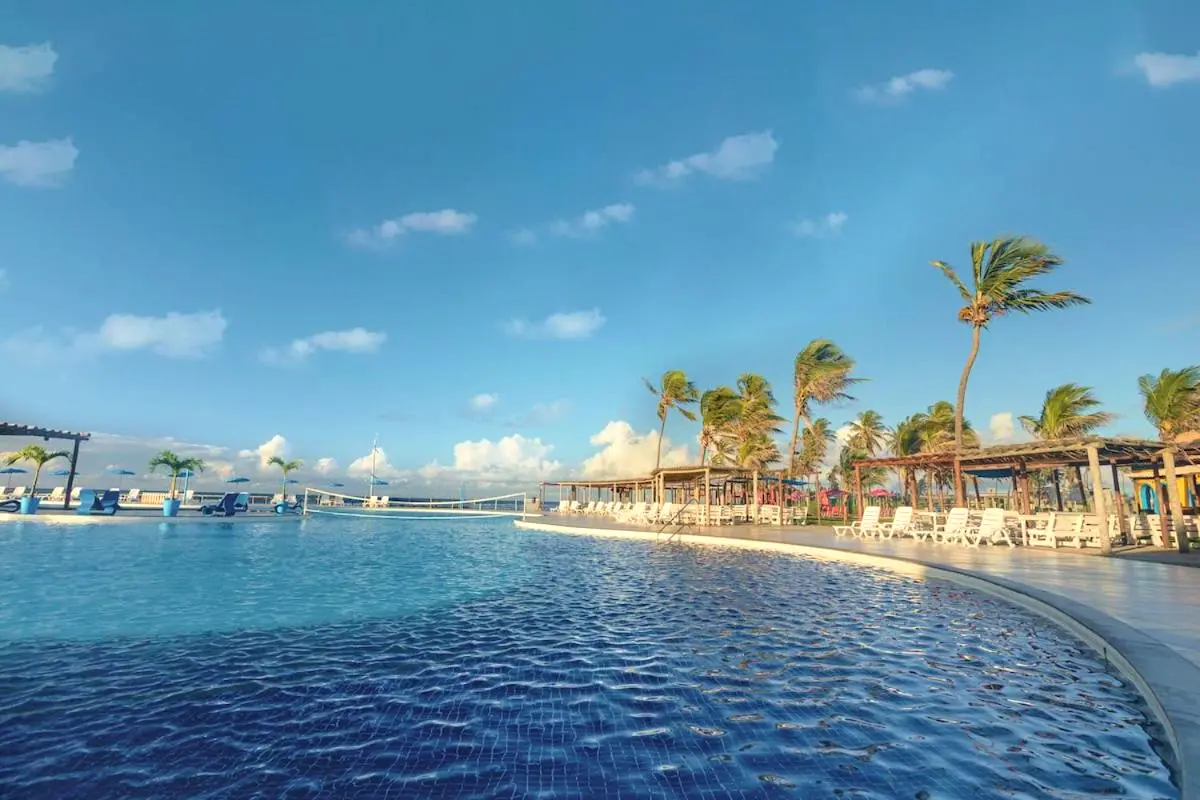 Piscina azul com palmeiras e quiosques de palha ao fundo