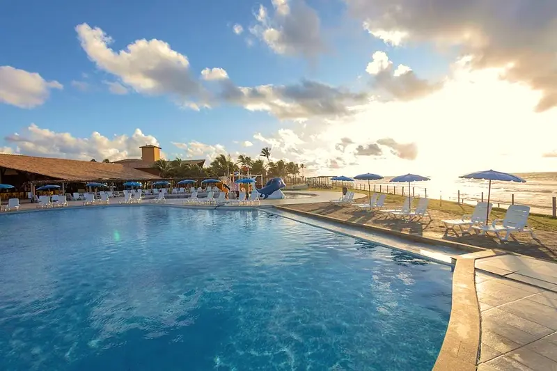 Piscina principal com espreguiçadeiras e área coberta de palha ao fundo